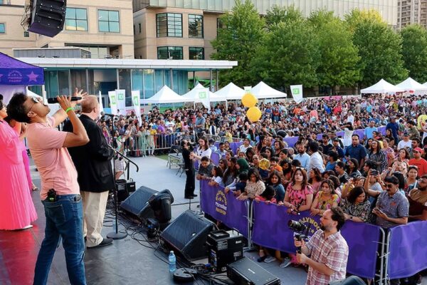An outdoor festival at Celebration Square. A performer to the left belts into a microphone in front of a crowd packed right up to a gate.