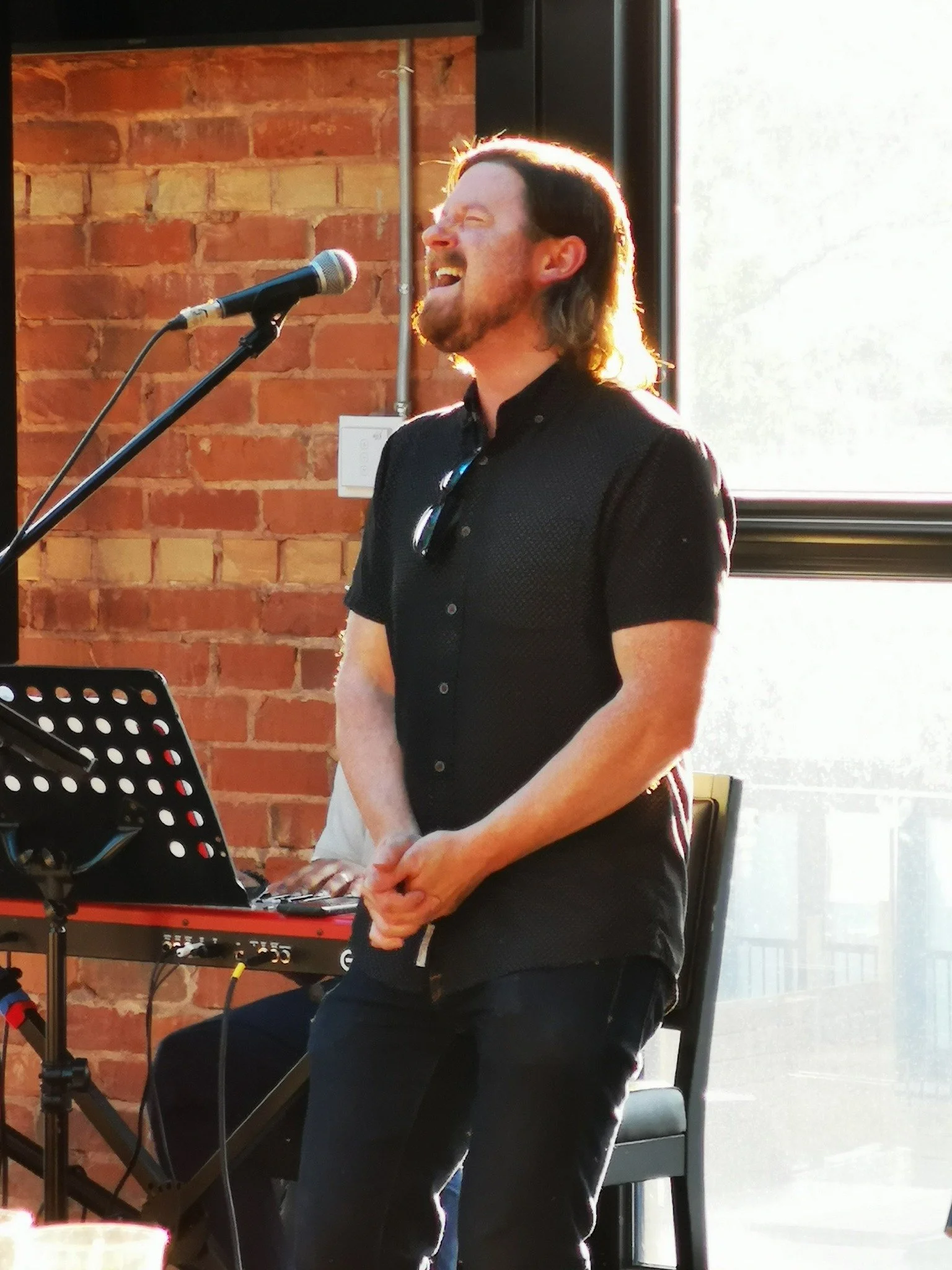 A man (Matt Zaddy) singing into a microphone in front of a brick wall. The hands and keyboard of a pianist can be seen behind him.