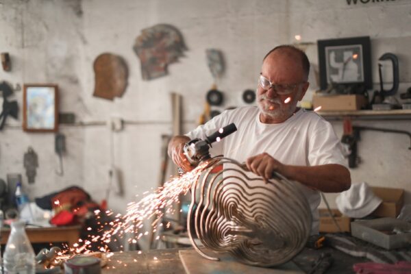 a Senior man using an angle grinder to create sculptures out of metal in his art studio.